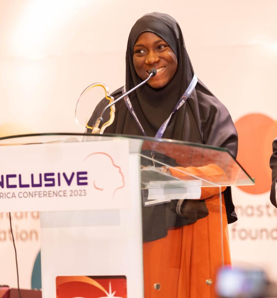 Anifat on a wine hijab with a microphone speaking at an event on disability inclusion.