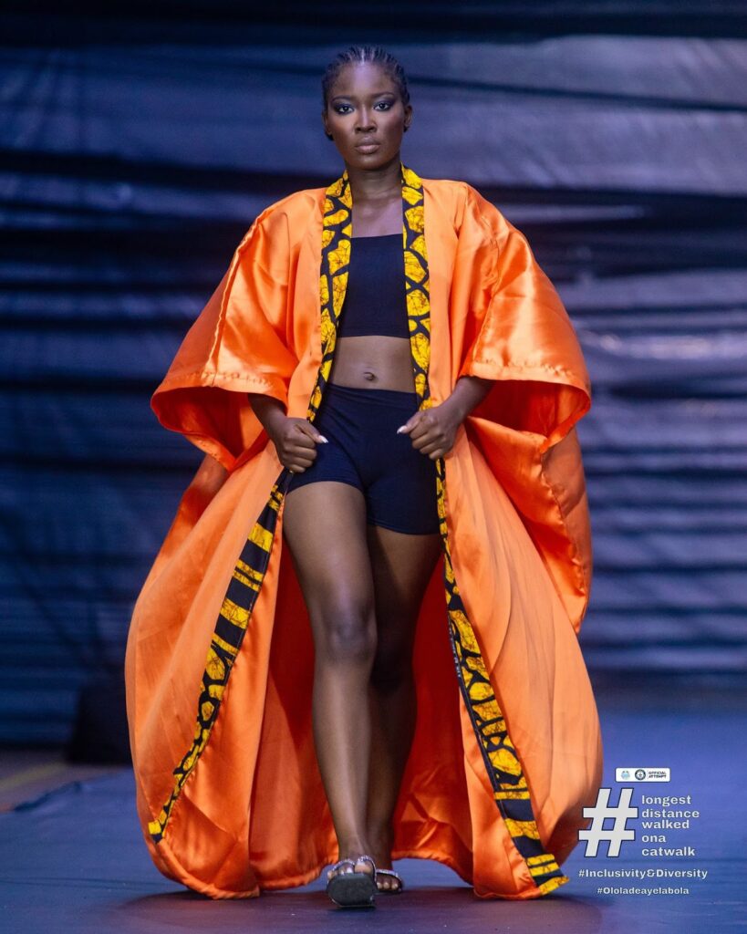 Model Ololade Ayelabola wears an orange overall jacket while walking on a runway